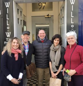 Five smiling people, two men and three women, stand in front a building entrance