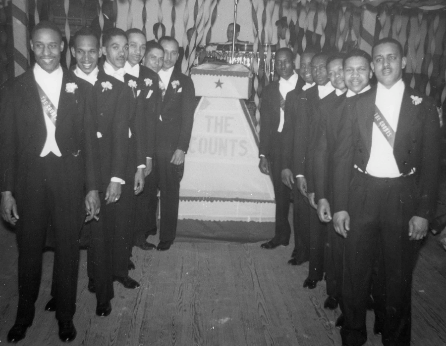 A group of twelve African American men from "The Counts" social club posing in formal tuxedos with tails and white gloves.